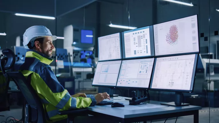 Industry 4.0 Modern Factory: Facility Operator Controls Workshop Production Line, Uses Computer with Screens Showing Complex UI of Machine Operation Processes, Controllers, Machinery Blueprints