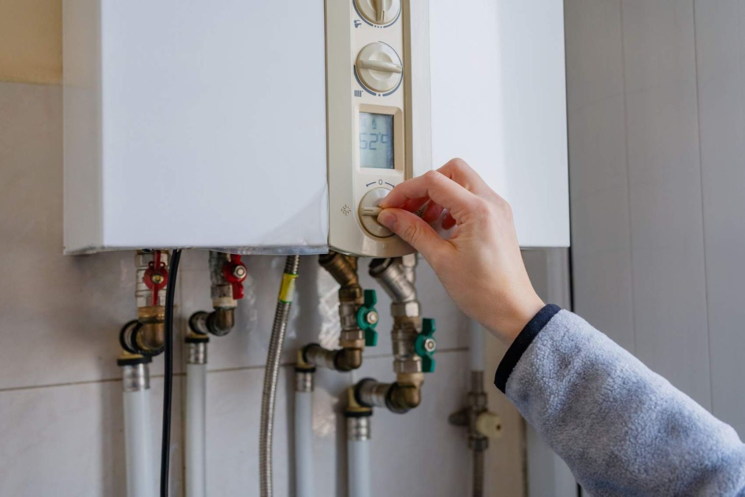 Hand turning the control knob on a white wall-mounted gas boiler, with pipes visible below, representing residential heating systems and everyday energy management at home.