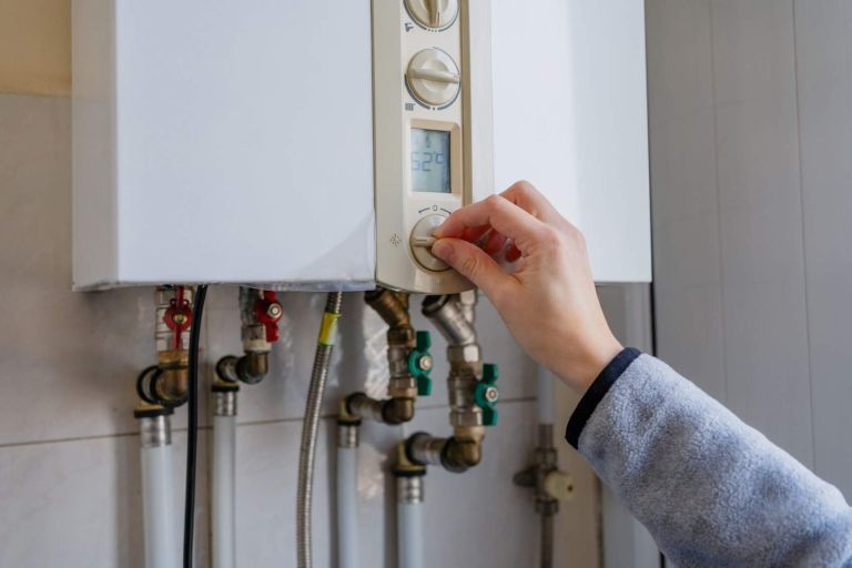 Hand turning the control knob on a white wall-mounted gas boiler, with pipes visible below, representing residential heating systems and everyday energy management at home.