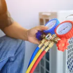 An air conditioner technician's hand holding a manifold gauge to check the pressure inside the system to normalize the refrigerant charge during high temperatures and hot weather. Maintenance concept.