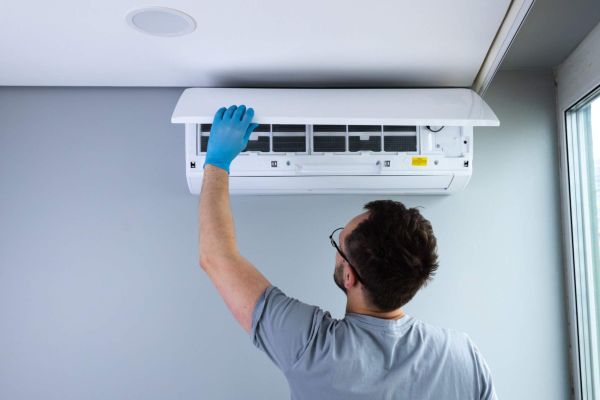 A worker in gloves is carefully checking and maintaining a ceiling-mounted air conditioning unit, ensuring its efficiency in a modern indoor environment with natural light flowing in.