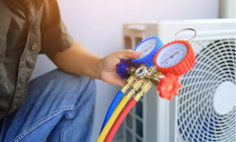 An air conditioner technician's hand holding a manifold gauge to check the pressure inside the system to normalize the refrigerant charge during high temperatures and hot weather. Maintenance concept.