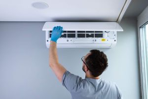 A worker in gloves is carefully checking and maintaining a ceiling-mounted air conditioning unit, ensuring its efficiency in a modern indoor environment with natural light flowing in.